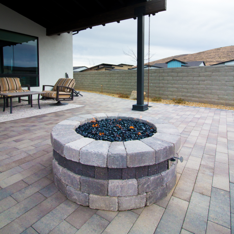 The Brown Residence boasts a backyard patio with a round stone fire pit filled with black lava rocks. Paved with beige and brown bricks, the patio includes cushioned outdoor chairs. A retaining wall encloses the yard, and a covered area is attached to the house.