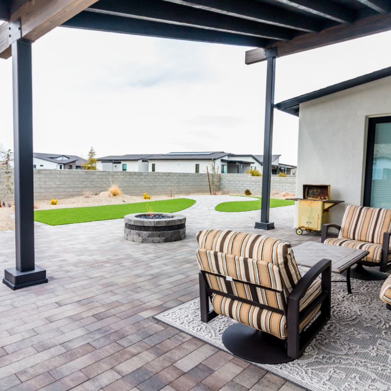 A modern backyard patio at the Brown Residence features striped cushioned chairs and a small yellow cart on a synthetic turf rug, overlooking a paved area with a circular fire pit. The covered patio is enclosed by a stone wall, offering views of neighboring houses.