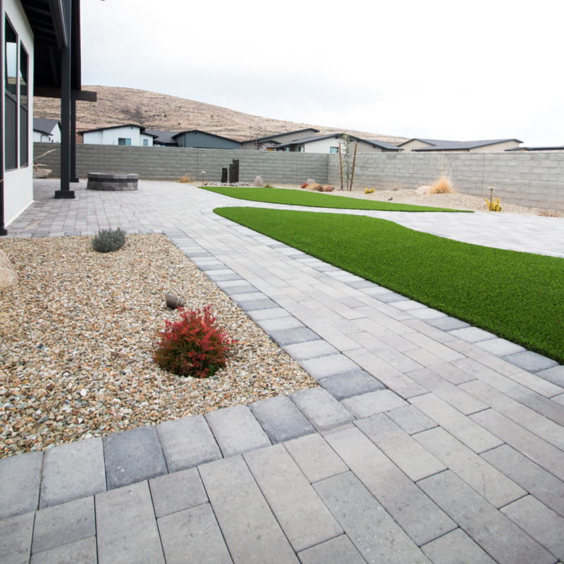 A modern backyard at the Brown Residence features a paved patio, artificial turf, and surrounding stone landscaping. This space includes a low concrete wall and is set against a backdrop of rolling hills and neighboring homes.