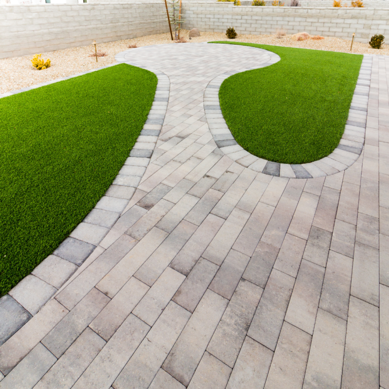 The Brown Residence boasts a neatly landscaped backyard featuring curved patches of green grass surrounded by light gray stone pavers, creating a modern and well-maintained appearance. The background has a stone wall and patches of decorative gravel with a few small plants.