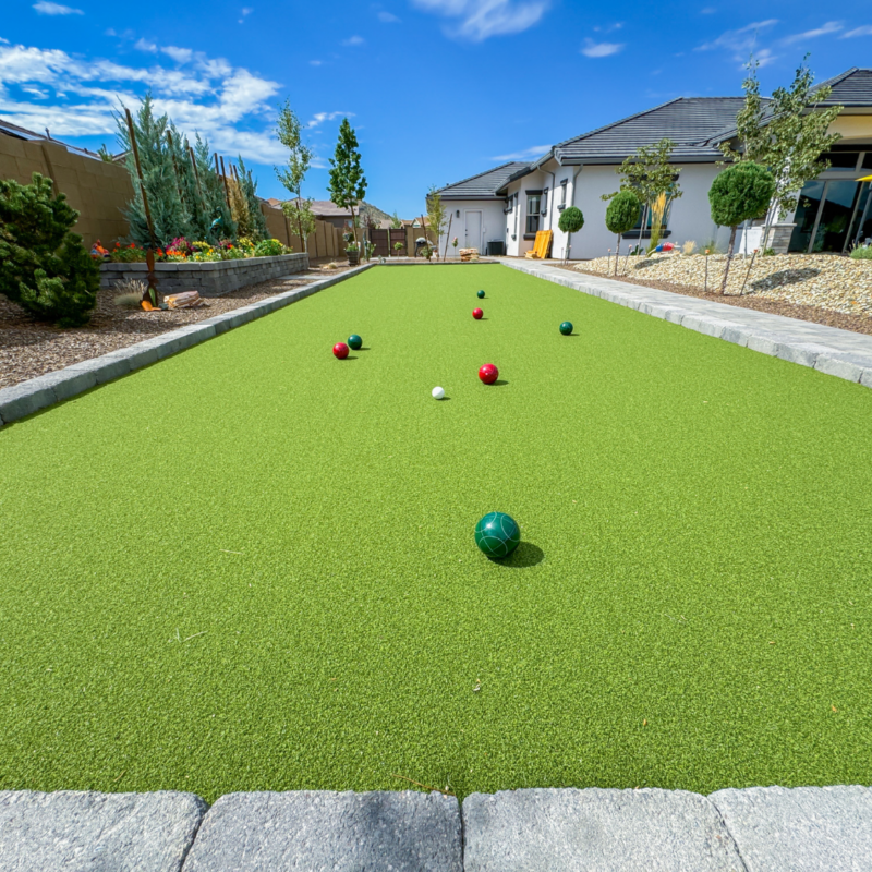 A residential backyard at the Hutchinson Residence featuring a bocce ball court with green turf. Several red and green bocce balls and a white pallino ball are scattered across the court. The yard is landscaped with trees, shrubs, and gravel beds. Houses and a clear blue sky are in the background.