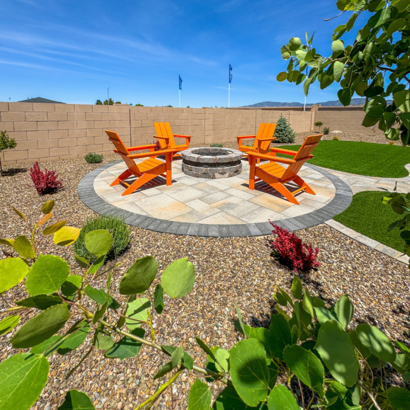 A backyard at Evermore Residence features a circular stone patio with a central fire pit and four bright orange Adirondack chairs. The yard is landscaped with gravel, lush greenery, red and green shrubs, and is surrounded by a tall stone wall. Clear blue sky in the background.