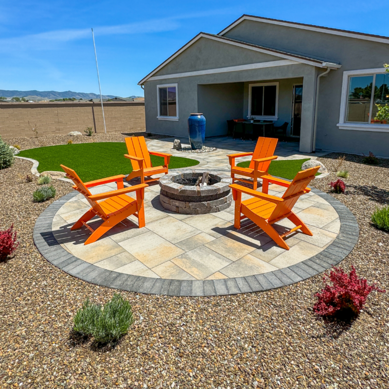 A sunny backyard scene at the Evermore Residence features a circular stone fire pit encircled by four bright orange Adirondack chairs on a paved patio. The yard boasts a green lawn, red and green shrubs, and a blue ceramic water feature near the charming single-story house.