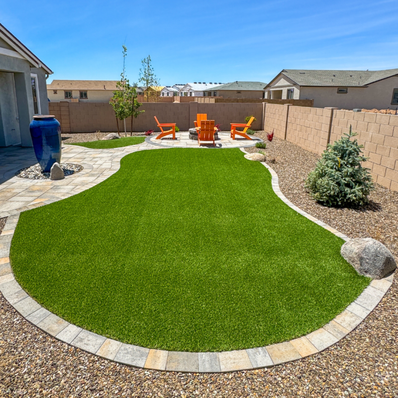 A backyard at Evermore Residence featuring a well-maintained lawn surrounded by a stone pathway and gravel. There&#039;s a cozy seating area with orange Adirondack chairs and a fire pit. A blue ceramic water feature stands to the left, and a small evergreen tree is near the right edge.