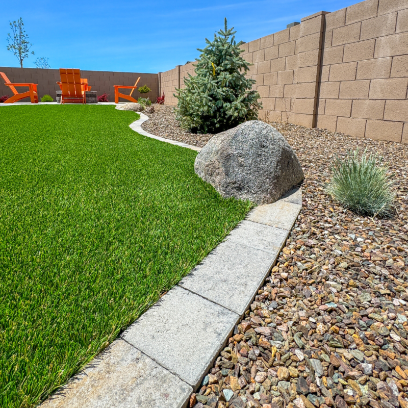 A manicured backyard at the Evermore residence features a lush green lawn bordered by a curved stone path. A large rock and small evergreen tree are placed amidst a gravel section. In the background, tan stone walls and bright orange outdoor chairs provide a pop of color under a clear blue sky.