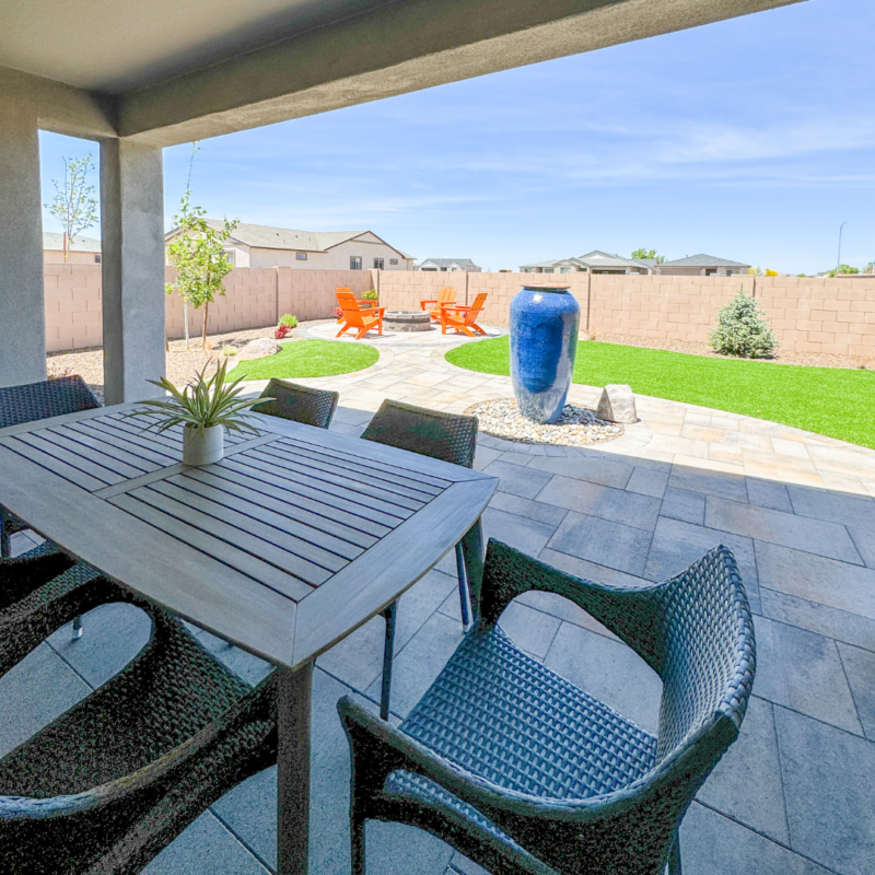 A covered patio with a dining table and four chairs overlooks the landscaped backyard at Evermore Residence. The yard features a grassy area, a large blue ceramic vase, and two bright orange Adirondack chairs. The sky is clear and blue, with houses visible beyond a brick wall.