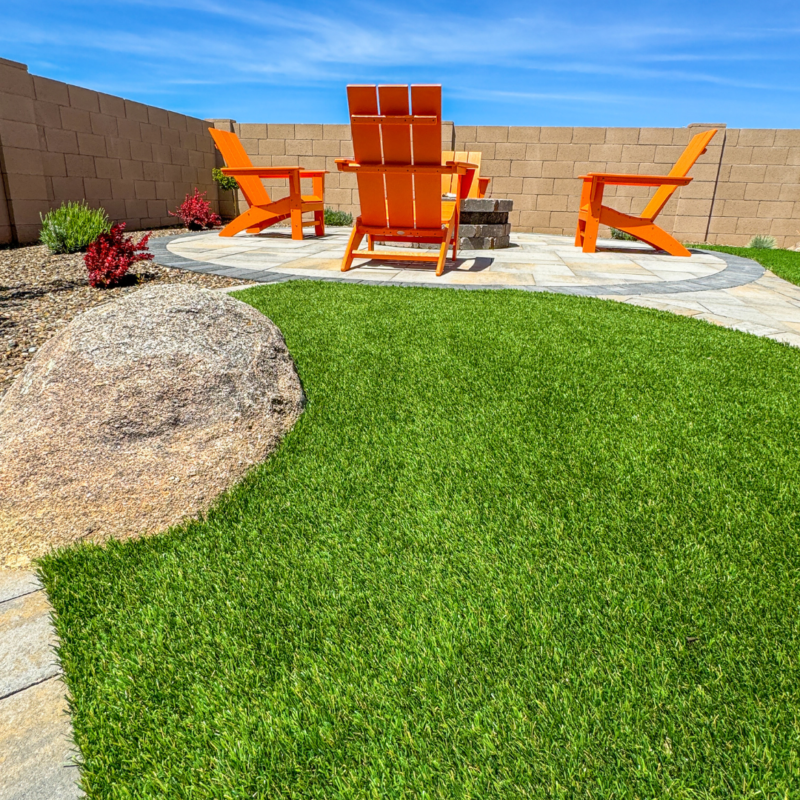 At the Evermore Residence, a backyard patio scene showcases vibrant green grass, a circular stone pathway, and three bright orange Adirondack chairs arranged around a small fire pit. The patio is bordered by a stone wall, with landscaping featuring desert plants and a large rock.