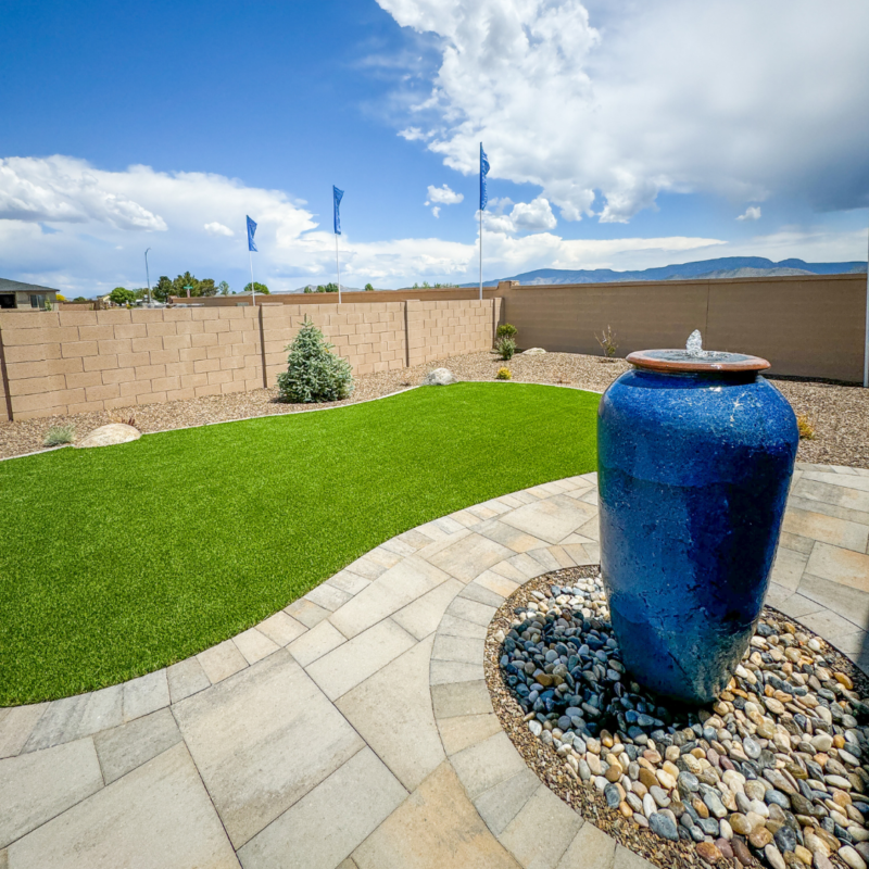 The Evermore Residence boasts a backyard with a lush green artificial lawn, stone tile patio, and a large blue ceramic water feature on a bed of rocks. A beige brick wall borders the yard, with several blue flags fluttering in the background under a partly cloudy sky.