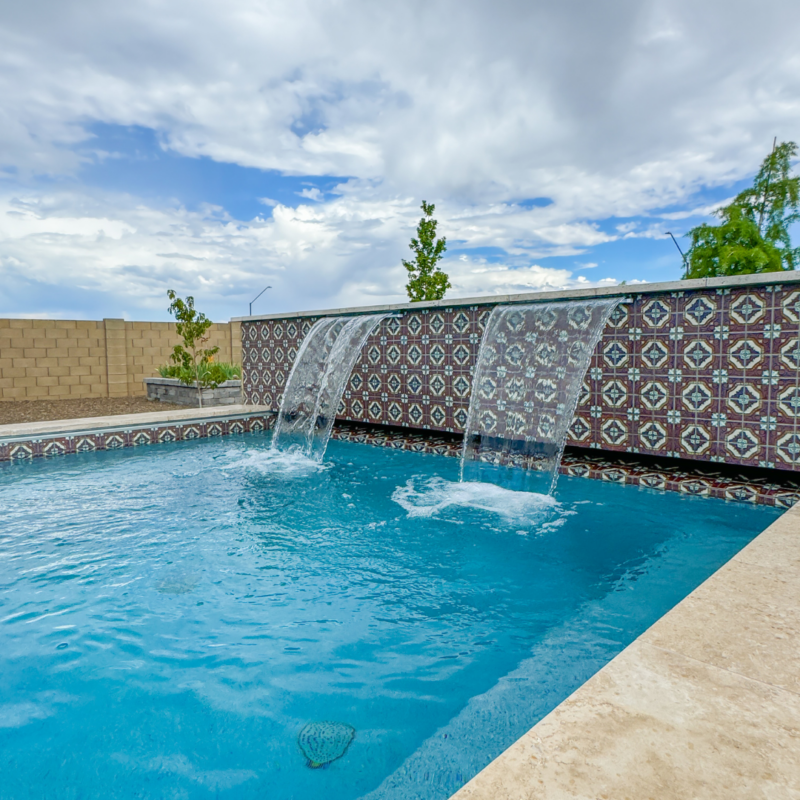 At the Hutchinson Residence, a modern backyard pool with decorative tile walls features two cascading waterfalls. The bright blue water contrasts beautifully with the intricate patterns of the tiles. The sky is partly cloudy, and there are some trees and a block wall fence in the background.