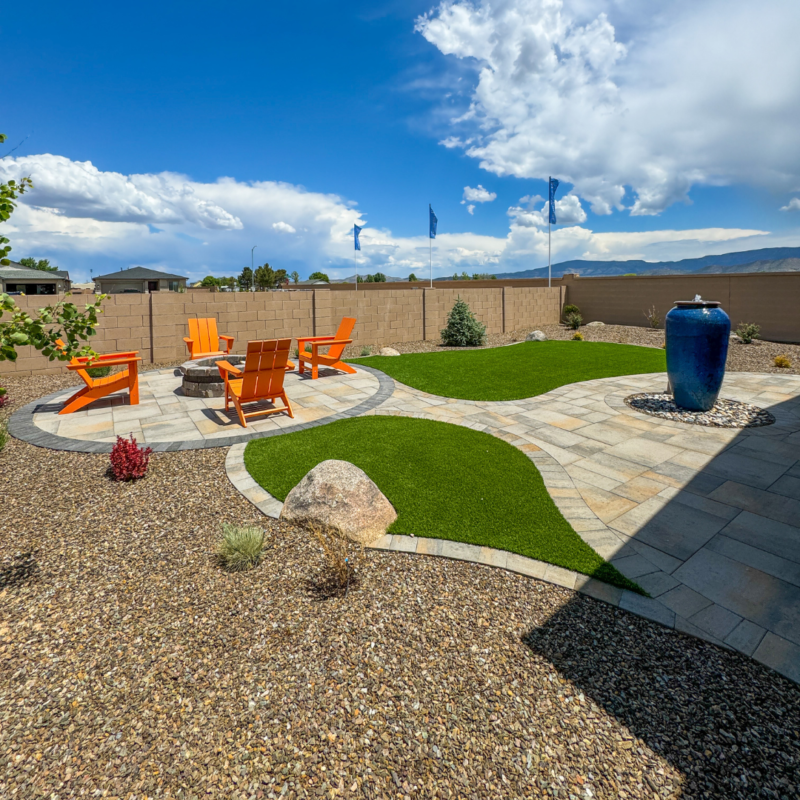 The Evermore Residence boasts a landscaped backyard with a circular patio featuring bright orange Adirondack chairs around a fire pit. The yard has a mix of artificial turf and gravel, a tall blue ceramic water feature, and a clear blue sky with some clouds overhead.