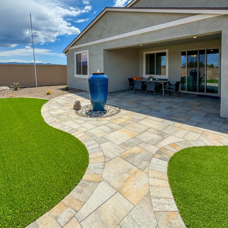 The Evermore Residence boasts a modern backyard with a tiled patio featuring a large blue vase as a focal point. A dining area with black chairs and a table is situated under a covered section of the patio. The yard is landscaped with artificial green turf and a stone pathway.