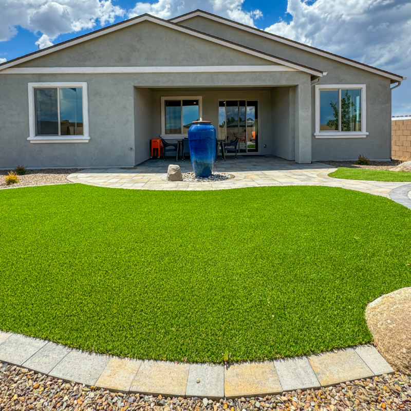 The Evermore Residence is a modern home with a manicured front yard featuring a lush green lawn and a circular stone pathway leading to the entrance. In the center of the yard is a large blue ceramic vase on a gravel patch. The house has light gray walls and large windows.