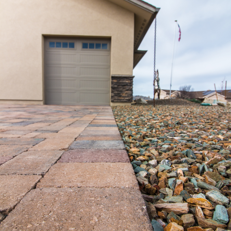 A close-up view of the Hodges Residence exterior shows a section of a paved driveway next to a landscaped area with small, multicolored rocks. The garage door is partially visible in the background, while an American flag flies proudly in the distance.