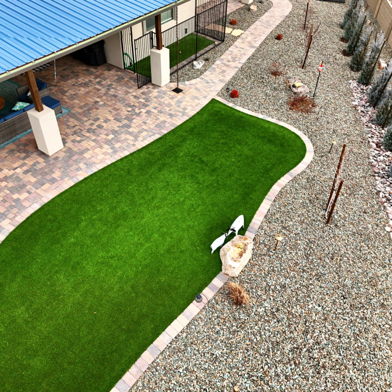 A well-maintained backyard at the Hodges Residence features a curvy patch of green artificial grass bordered by bricks. The surrounding landscape includes gravel and a few sparse plants. A paved patio with a blue roof is visible on the left side, accompanied by some outdoor furniture.