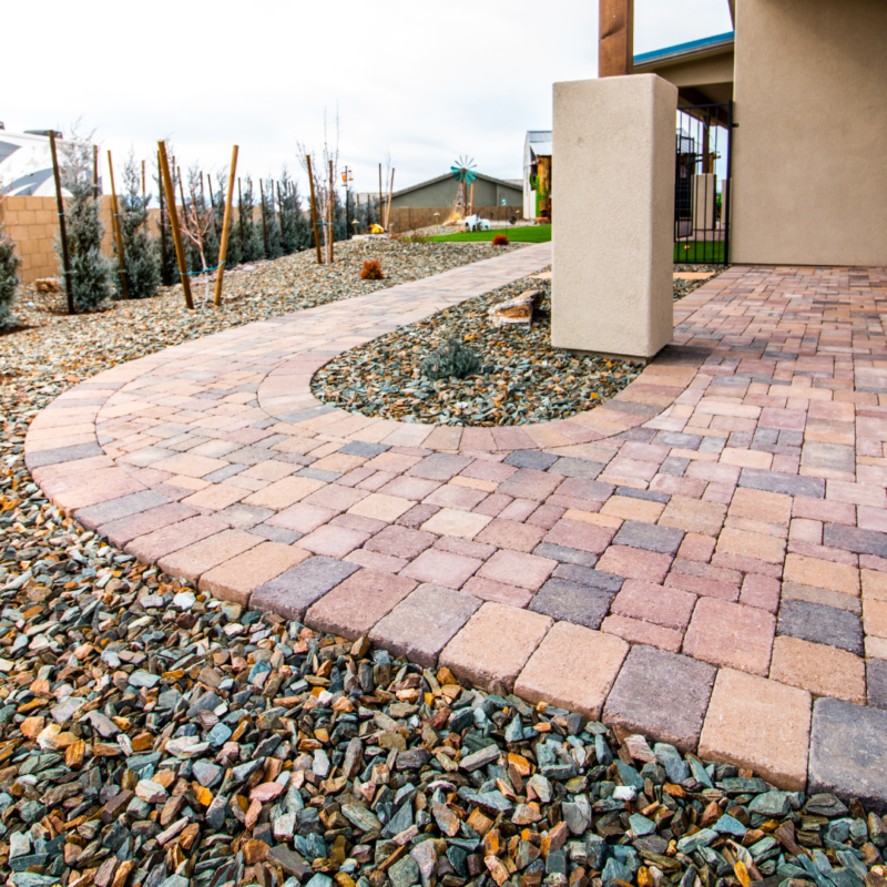 A landscaped yard at the Hodges Residence features a curved pathway made of interlocking brick pavers. The path is bordered by decorative rocks and leads to a modern building. Young trees and shrubs line the yard, and a windmill is visible in the background.