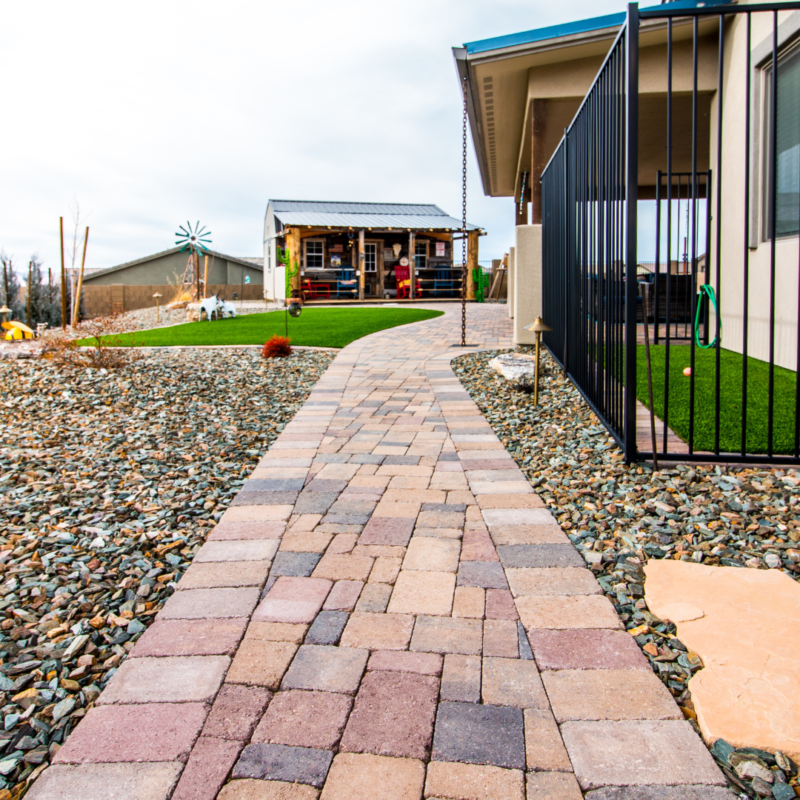 A stone pathway leads through the Hodges Residence yard with rocks and sparse vegetation, bordered by a black metal fence on the right and a building. In the background, a small structure of wood and glass has people near it, with a windmill visible to the left.