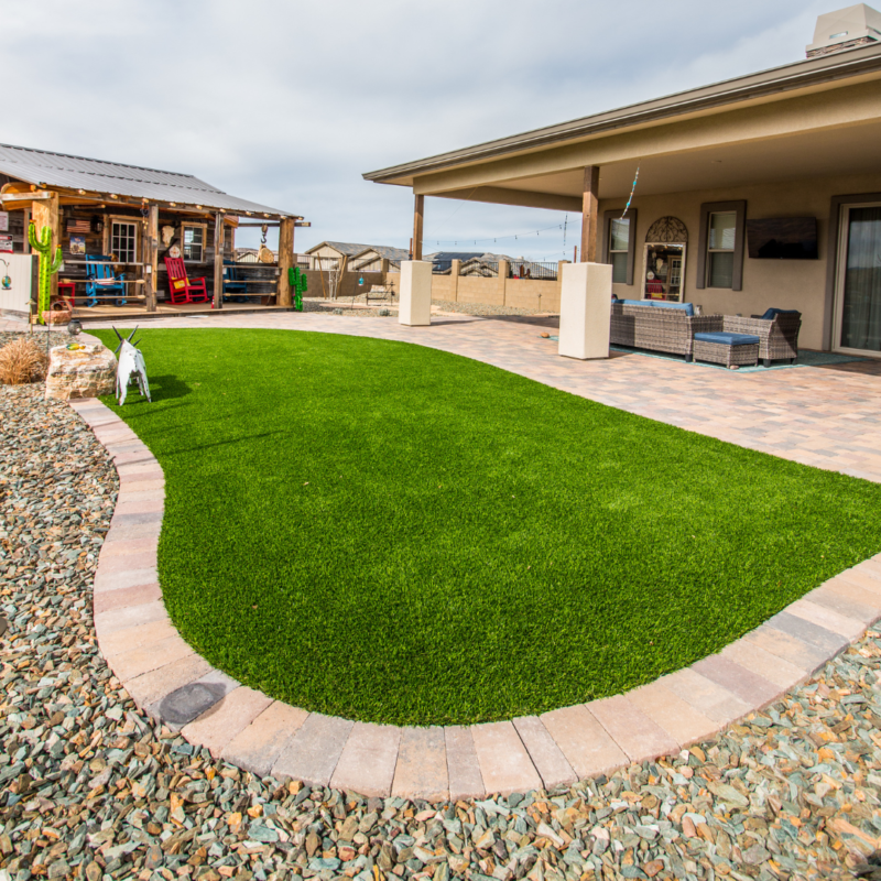 At the Hodges Residence, a well-maintained backyard boasts a neatly trimmed green lawn surrounded by stone pavers. The yard features a cozy patio with outdoor furniture and a wooden structure in the background. A small decorative windmill and rocky landscaping are also visible.