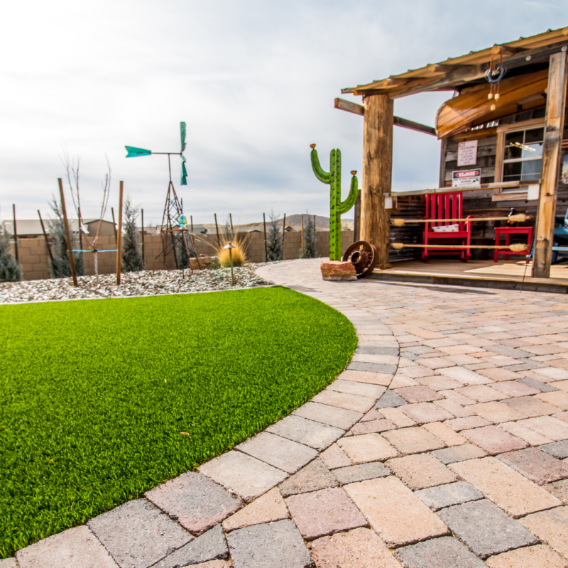 The Hodges Residence boasts a landscaped backyard featuring a curved brick pathway leading to a wooden structure with a red rocking chair. A fake green lawn sits beside the path, complemented by decorative elements like a windmill, a large cactus statue, and various outdoor decor items.