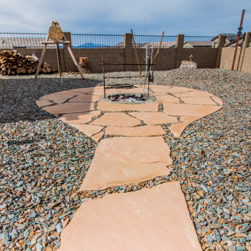 The Hodges Residence boasts a backyard with a stone pathway leading to a circular fire pit area paved with large, irregularly shaped stones. The yard is covered in gravel, with stacks of firewood in the background and a fence and mountains visible in the distance.