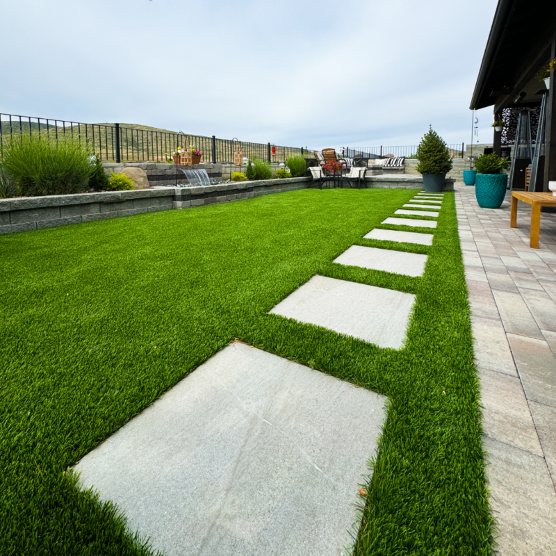 The Bauman Residence features a backyard with a well-manicured green lawn and large square stepping stones leading to a seating area. The space is bordered by a stone retaining wall and decorative plants. In the background, there&#039;s a patio with outdoor furniture and a scenic view beyond.