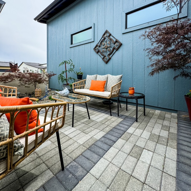 At the Bauman Residence, a cozy outdoor patio with wicker furniture adorned with orange and white cushions invites relaxation. The patio is paved with grey and black tiles and features a blue wall with two narrow horizontal windows and decorative wall art. Potted plants add greenery to the scene.