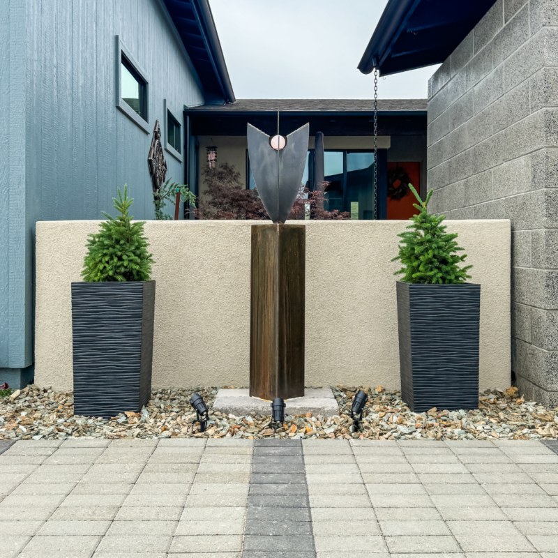 A modern outdoor sculpture is flanked by two tall potted plants in a small courtyard at the Bauman Residence, with beige walls and contemporary buildings on either side. The area features a paved walkway and decorative rocks surrounding the planters.