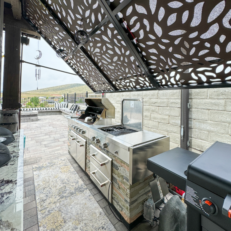 The Bauman Residence outdoor kitchen setup features a stainless steel built-in grill and stovetop, under a decorative leaf-patterned metal pergola. The area includes countertop space and a view of the patio with hills in the background. The surface and structure are modern and well-maintained.