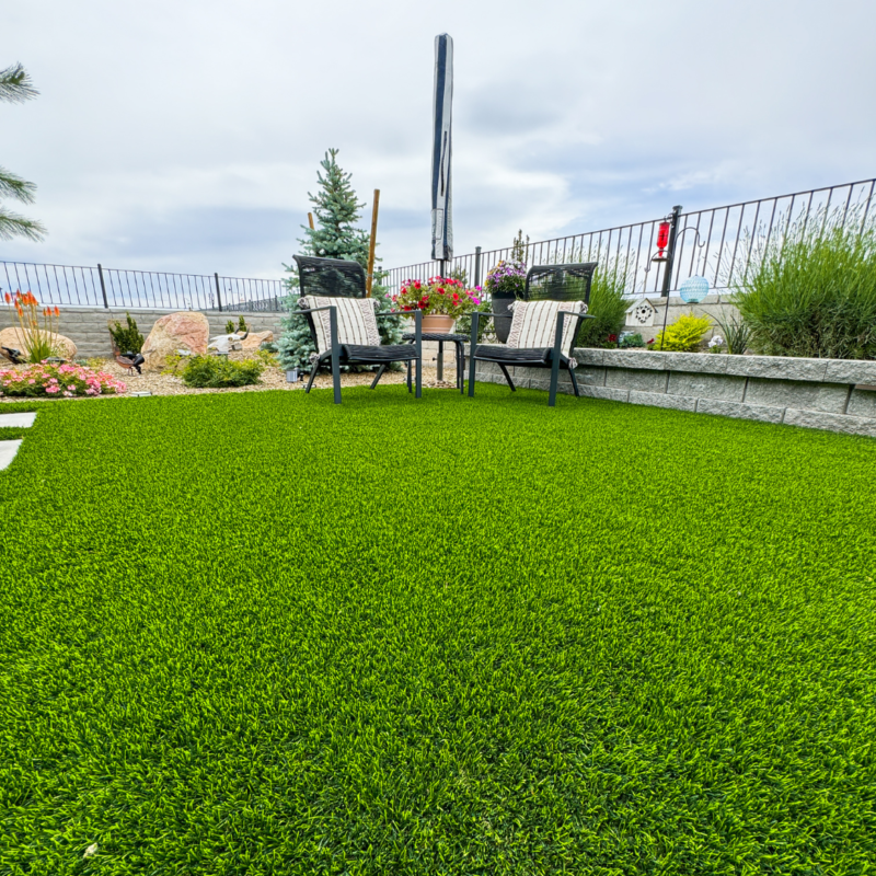 The Bauman Residence boasts a neatly manicured lawn with a small outdoor seating area consisting of two chairs and a table adorned with flowers. The background features a well-maintained garden, a fence, and a partly cloudy sky.