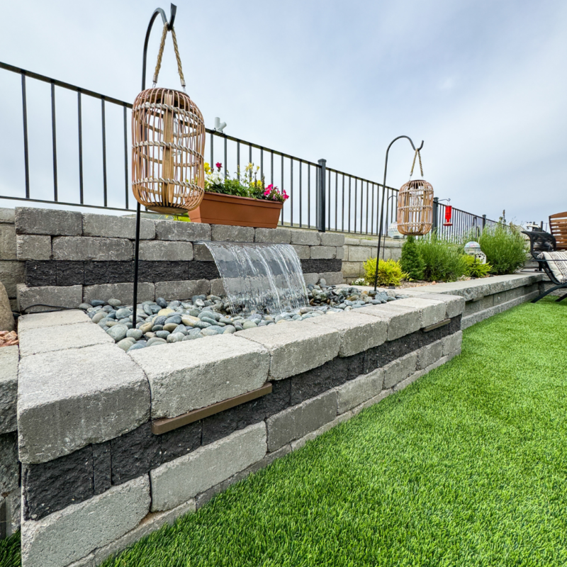 The Bauman Residence boasts a stone-tiered garden with a small waterfall feature. Two wicker hanging baskets frame the waterfall, which is surrounded by smooth stones. Above the waterfall is a planter box with flowers. The garden includes a lush, green lawn and a black metal fence in the background.