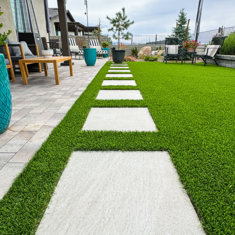 The Bauman Residence boasts a backyard with a neatly mowed lawn and large rectangular stone pavers leading to a seating area. The patio features wooden furniture with cushions, potted plants, and a few small trees. Enclosed by a metal fence, the area complements the partly cloudy sky perfectly.