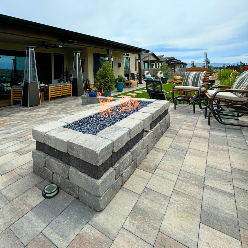 A patio at the Bauman residence features a brick fire pit with flames burning, surrounded by outdoor furniture including cushioned chairs. The area is paved with stone tiles, and a well-maintained garden with various plants and decorations lines the perimeter of the house in the background.