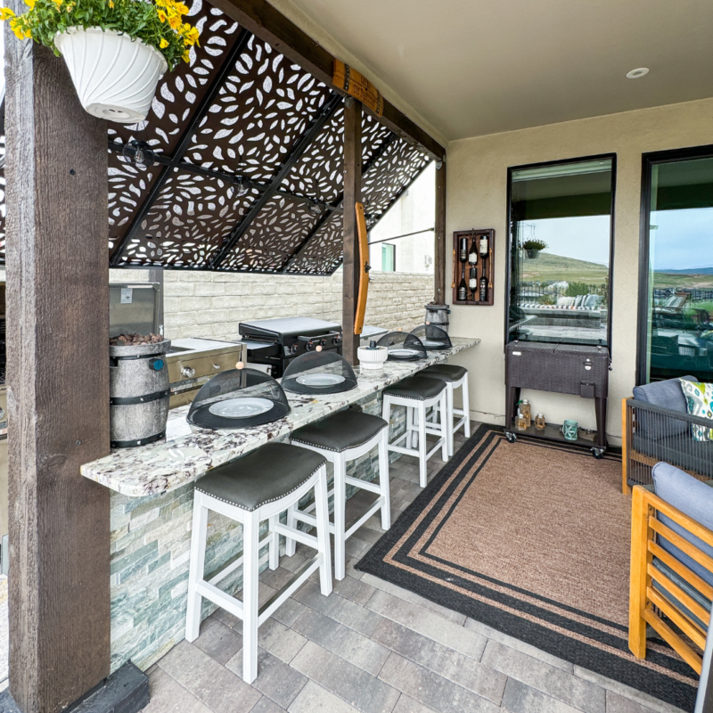 Outdoor kitchen at the Bauman Residence with a patterned pergola roof, granite countertops, and four white bar stools. Features a grill, cooking utensils, a hanging plant, and various cooking supplies. Adjacent to a cozy seating area with an outdoor rug and scenic view in the background.