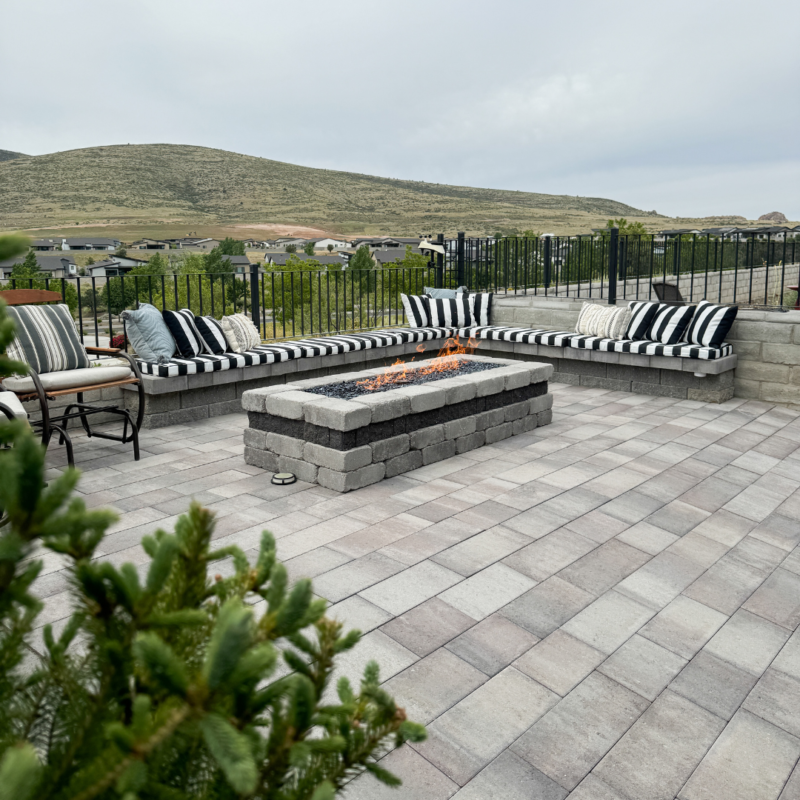 The Bauman Residence features an inviting outdoor patio with a stone fire pit at its center, surrounded by cushioned benches adorned with black-and-white striped pillows. Behind the patio, a sleek black metal fence offers views of a grassy hillside with houses in the distance, framed by pine branches in the foreground.