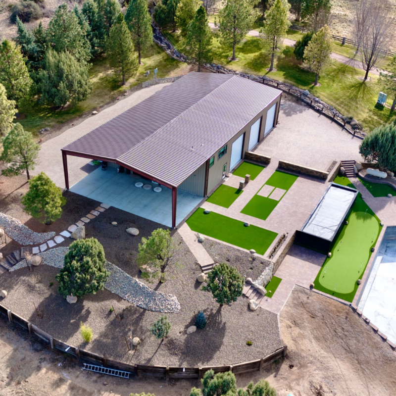 Auto Draft: Aerial view of a modern, rectangular metal building with a covered patio, surrounded by landscaped areas with trees, rocks, and gravel pathways, various green lawns, a putting green, and adjacent paved areas within a spacious, open setting.