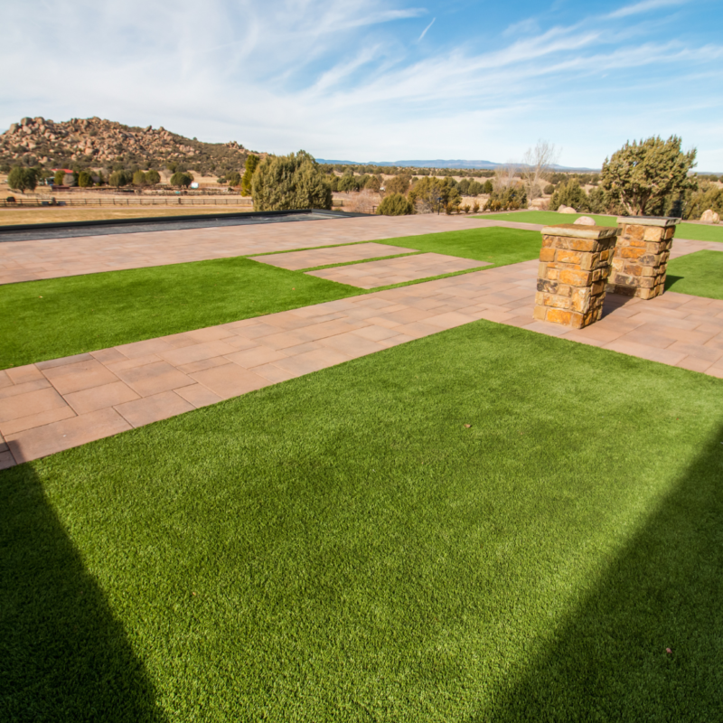 Outdoor backyard landscape featuring a large artificial green lawn bordered by stone pathways and rectangular planters. Auto Draft selections enhance the backdrop of trees, shrubs, stone wall, and hills under a blue sky with scattered clouds.