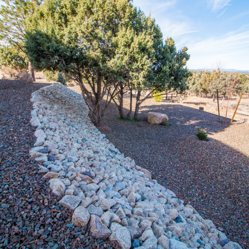 A dry landscape with a slope reinforced by white stones, leading to a rocky area with scattered shrubs and trees. In the background, there&#039;s a wooden fence and open land extending towards the horizon under a clear blue sky—an auto draft of nature’s raw beauty.