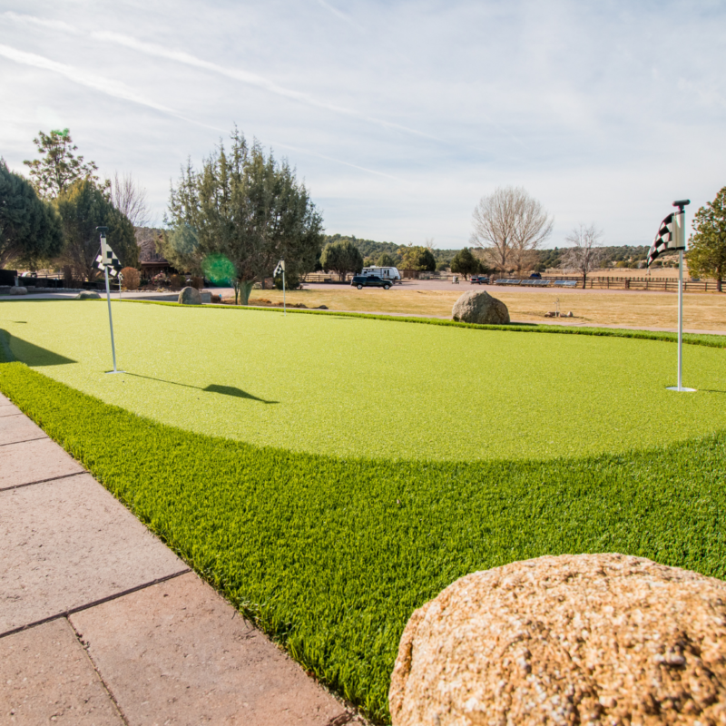 A bright green golf putting green with smooth artificial turf is surrounded by landscaping rocks and a stone pathway. In the background, trees and shrubs dot the landscape beyond the Brundy residence, where a car is parked under a clear, sunny sky.