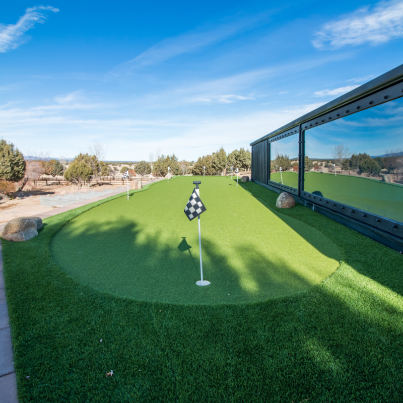 A small, well-manicured putting green with a checkered flag on a sunny day. The green is bordered by rocks and surrounded by lush grass. In the background, there are trees and a clear, blue sky with a few wispy clouds—an auto draft of nature&#039;s perfection.