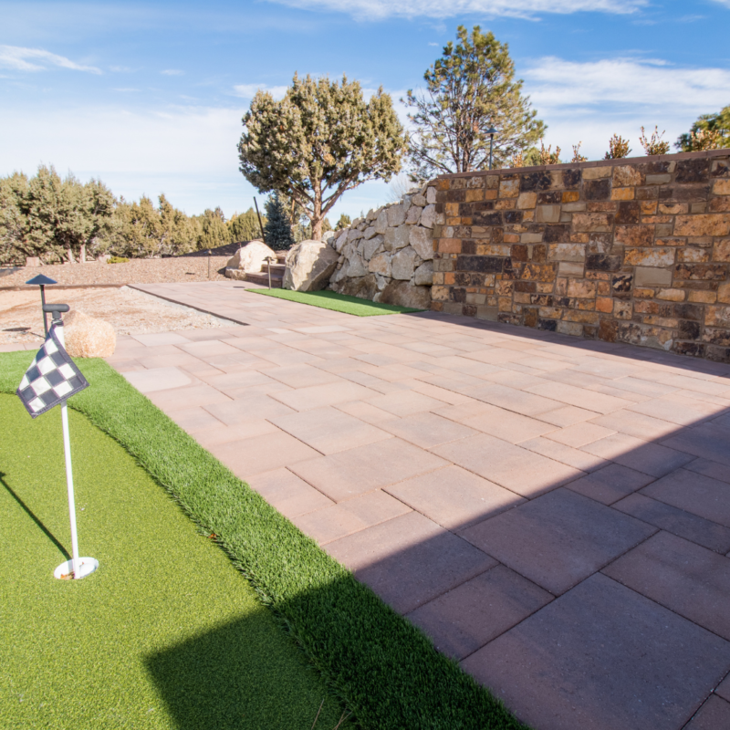 A serene outdoor scene featuring a small putting green with a checkered flag, adjacent to a tan and brown brick patio. Trees, shrubs, and a stone wall are in the background under a blue sky with wispy clouds. A large rock is visible near the edge of the patio, creating an idyllic auto draft setting.