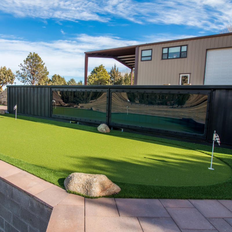 A modern building with a garage is in the background, and a sleek rectangular swimming pool with dark glass siding is in the foreground. The pool is surrounded by artificial turf with two small flags marking holes, indicating a mini-golf area, reflecting an impeccable auto draft design aesthetic.