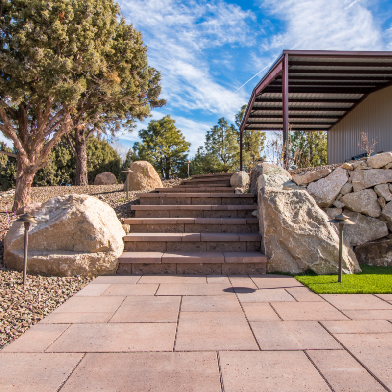 A landscaped outdoor area with stone steps leading up to a metal-roofed structure. Large rocks and trees flank the steps, and a paved area with artificial grass is in the foreground. The sky is clear with scattered clouds, creating a scene that looks as if it were created by Auto Draft.