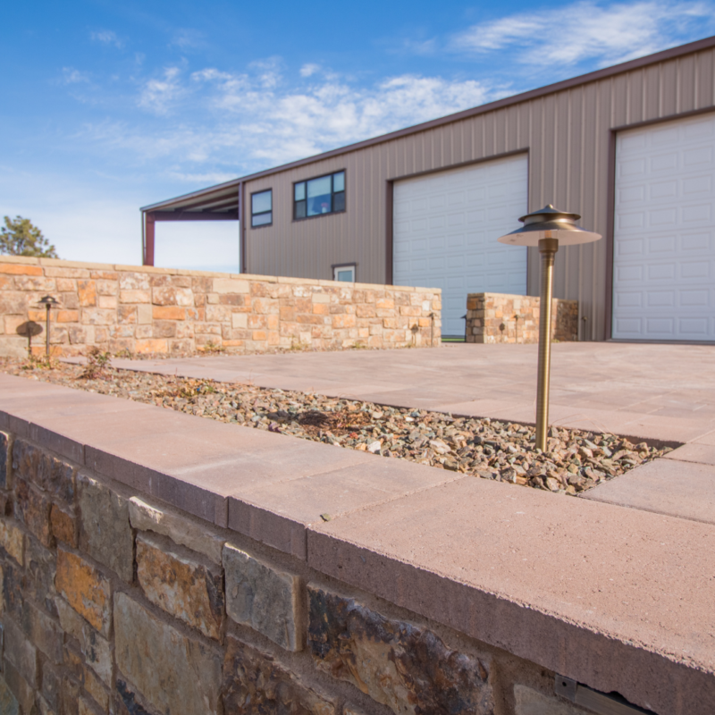 A modern industrial building with large white garage doors and tan siding, designed perfectly for auto draft services. The structure is surrounded by a brick and stone retaining wall, while small gravel beds and a solitary metal lantern add detail to the outdoor space. The sky is clear and blue.