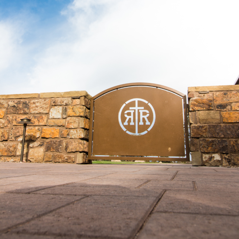 A brown metal gate set in a stone wall features a logo with interlocking letters &quot;RT&quot; and a cross in the center. The ground is paved with square bricks, resembling an auto draft design, and the sky is bright with clouds.