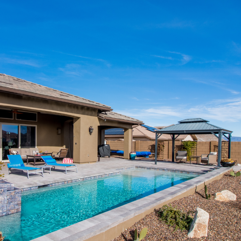 The Hutchinson Residence features a modern house with a backyard showcasing a clear swimming pool, patio furniture including lounge chairs with cushions, a grill, and a gazebo. The yard is meticulously landscaped with rocks and plants, all set under a bright blue sky.