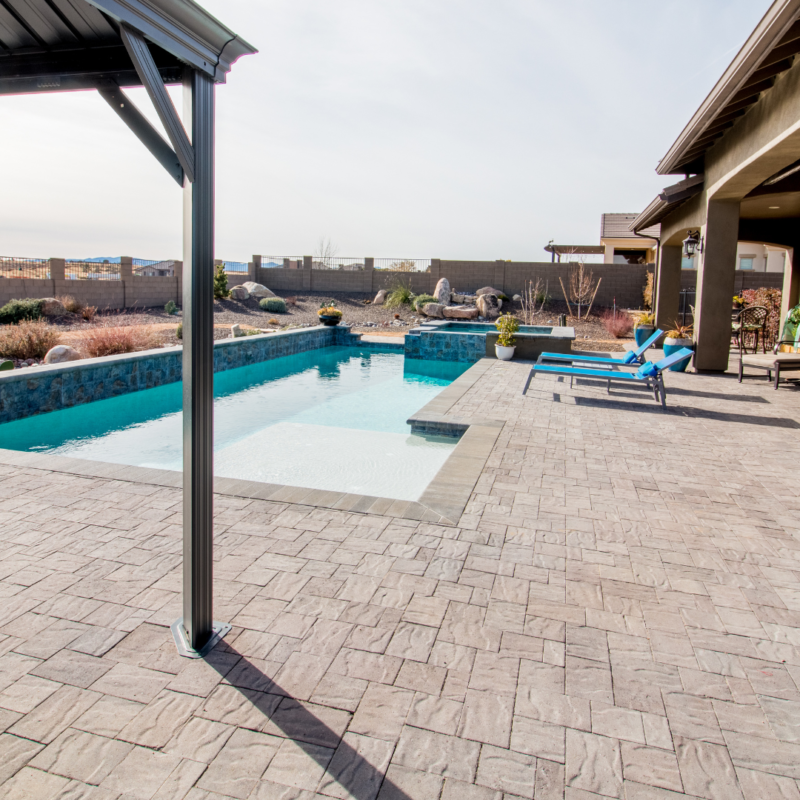 At the Hutchinson Residence, the backyard features a rectangular swimming pool with a shallow lounging area and steps leading into the main pool. Two blue lounge chairs sit on the stone-paved poolside. A covered patio with seating is visible on the right, and a stone wall encloses the yard.