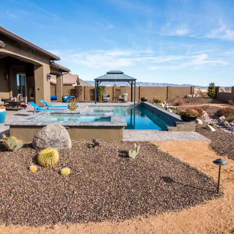 A backyard scene at the Hutchinson Residence features a rectangular swimming pool with a stone deck. There is a gazebo at the far end of the pool and a house to the left. The surrounding landscape includes desert plants and cacti, with a mountain range visible in the distance under a blue sky.