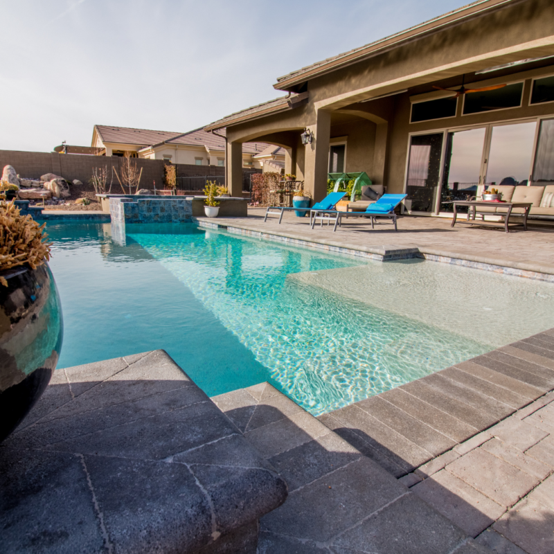 A backyard at the Hutchinson Residence features a modern swimming pool with crystal-clear water, surrounded by a gray stone patio. Blue lounge chairs and a shaded seating area under a covered patio offer relaxation. In the background, neighboring residences are visible under a blue sky.