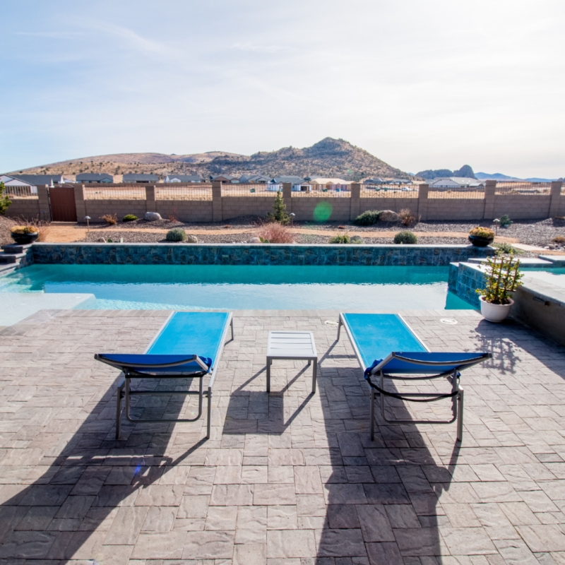 The Hutchinson Residence features a backyard with a swimming pool, two blue lounge chairs, and a small table between them. The area is surrounded by a stone patio, desert landscaping, and a fence. Mountains rise in the background under a clear sky.