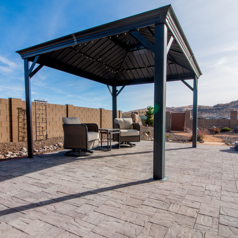 A backyard scene at the Hutchinson residence shows a covered patio structure with a metal roof. Underneath, two cushioned chairs with a small table between them are placed on a stone-paved surface. The area is surrounded by a stone wall, and a mountainous landscape is visible in the background.