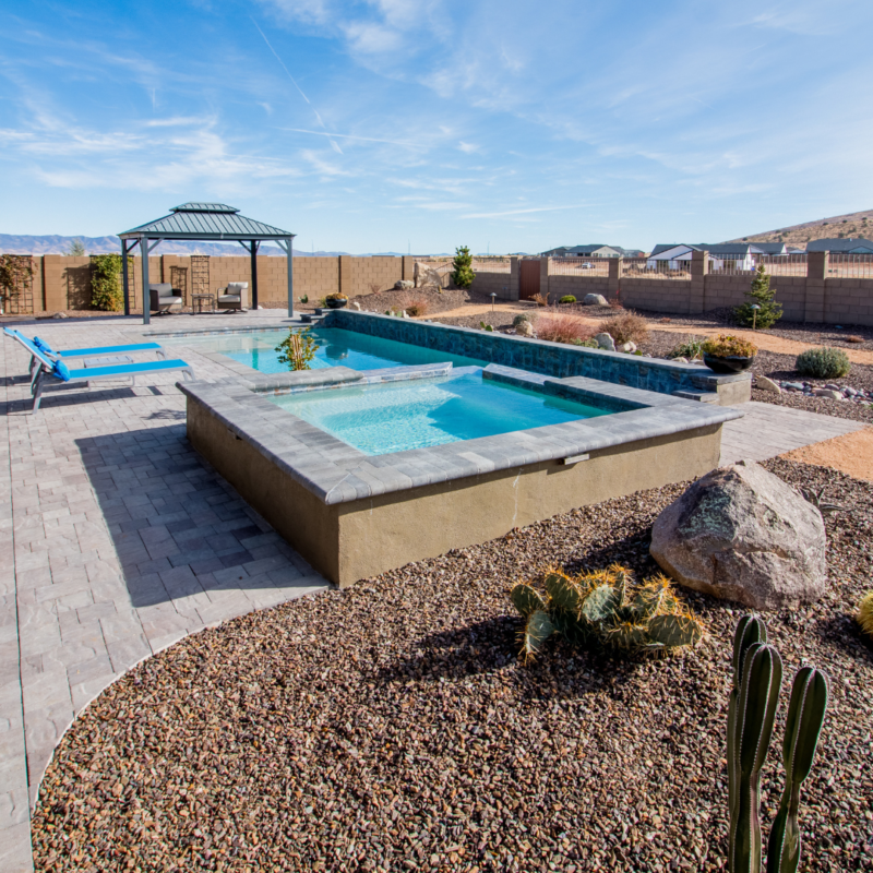 The Hutchinson Residence features a backyard with a rectangular swimming pool elevated by stone walls, surrounded by a stone patio. A small gazebo sits in the background next to a pool lounger. The landscape includes desert plants and rocks, all enclosed by a wooden fence.